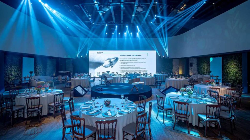 Empresa de eventos: o que observar antes de escolher o espaço para seu evento retrata um ambiente sofisticado e bem estruturado, ideal para eventos corporativos. O espaço conta com mesas elegantemente decoradas, dispostas em formato circular, cada uma arrumada com louças refinadas, taças e arranjos florais.