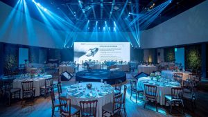 retrata um ambiente sofisticado e bem estruturado, ideal para eventos corporativos. O espaço conta com mesas elegantemente decoradas, dispostas em formato circular, cada uma arrumada com louças refinadas, taças e arranjos florais.