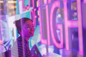 A imagem retrata perfeitamente o conceito de brand experience. Nela, uma jovem observa, encantada, uma vitrine iluminada com luzes de néon vibrantes em tons de rosa, roxo e azul.