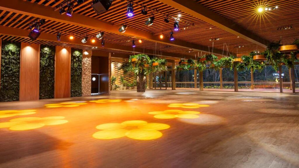 Casa de show moderna com piso de madeira, iluminação cênica e plantas suspensas, preparada para eventos corporativos com palco e estrutura de som profissional.