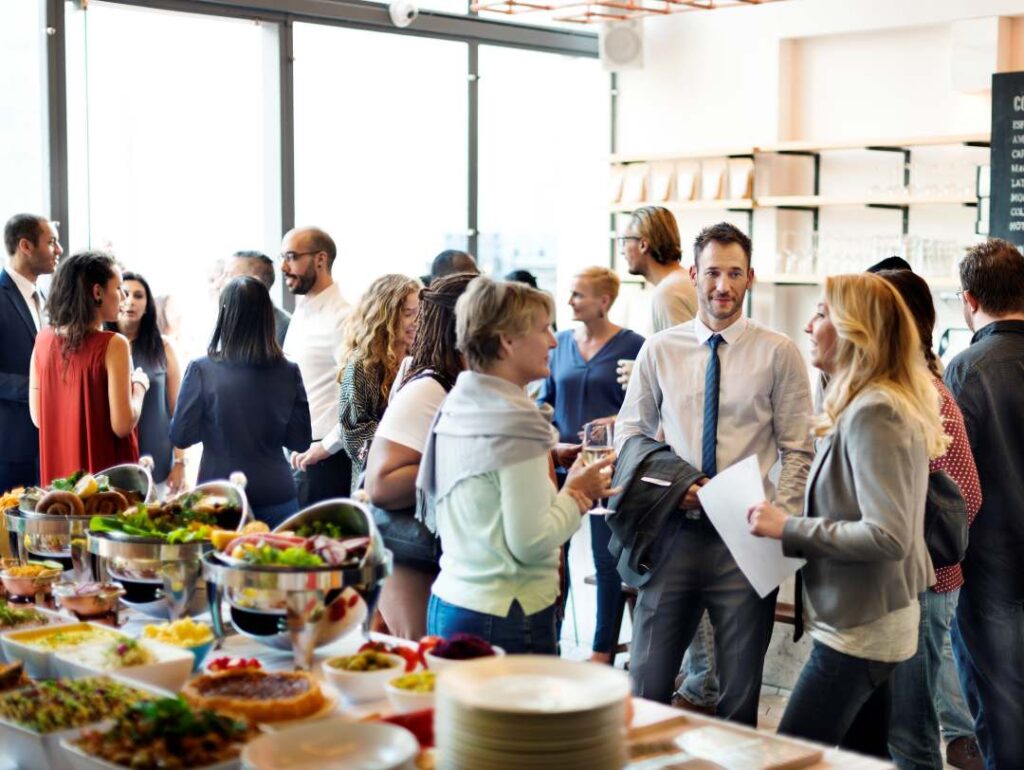 Como organizar um evento corporativo de sucesso: do planejamento à execução impecável grupo de pessoas interagindo em um ambiente moderno e bem iluminado, ao lado de uma mesa repleta de opções gastronômicas.