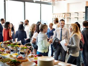 grupo de pessoas interagindo em um ambiente moderno e bem iluminado, ao lado de uma mesa repleta de opções gastronômicas.