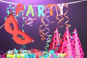 Em frente a um fundo roxo, a palavra "PARTY" feita de letras coloridas está pendurada com fitas. Abaixo, uma máscara vermelha decorada com estrelas brilhantes flutua no ar. No canto direito, três chapéus de festa rosa com bolinhas pretas estão apoiados juntos.