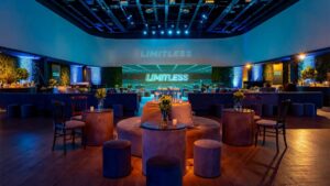 A imagem retrata uma sofisticada decoração de festa em um ambiente corporativo moderno e imersivo. O espaço é amplo, com iluminação cênica em tons de azul que valorizam cada elemento do cenário. O palco ao fundo destaca a palavra “LIMITLESS” em uma projeção tecnológica, reforçando o conceito do evento. A disposição dos móveis, mesas redondas baixas com banquetas e poltronas, cria áreas de convivência elegantes e acolhedoras. Elementos naturais, como painéis verdes nas laterais, trazem um toque orgânico ao design contemporâneo. Essa decoração de festa une tecnologia, conforto e estética para oferecer uma experiência visual impactante e memorável.