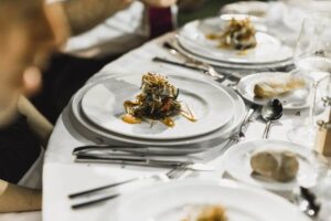 Imagem de uma mesa elegantemente posta durante um evento, com pratos de alta gastronomia servidos individualmente. Ao centro dos pratos brancos, há uma montagem refinada de ingredientes cuidadosamente dispostos, com molhos decorativos e finalização gourmet. Talheres de inox, taças de vidro e pães artesanais complementam o cenário. O ambiente transmite sofisticação e atenção aos detalhes, característicos de jantares de alto padrão.